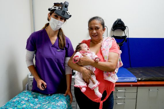 Eine Frau im rosa Kttel steht neben einer anderen, die ein Baby auf dem Arm hält.
