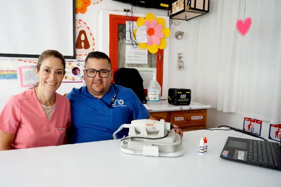 Eine Frau im rosa Kttel und ein Mann im blauen Kittel sitzen an einem Tisch. Darauf stehen ein Telefon mit Hörer und ein Computer mit Tastatur.