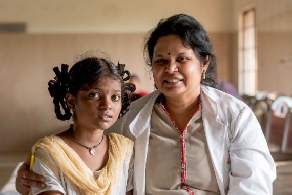 Eine Frau im weißen Kittel sitzt neben einem Mädchen, um dessen Schultern sie ihren Arm gelegt hat.
