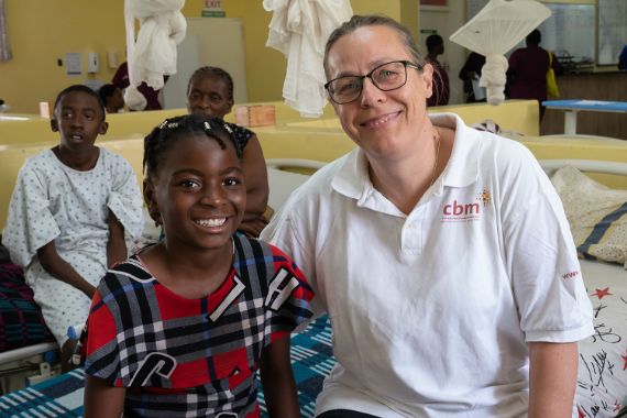 Eine Frau im weißen Poloshirt mit CBM-Logo sitzt neben einem etwa zehnjährigen Mädchen auf einem Bett in einem Patientenzimmer.