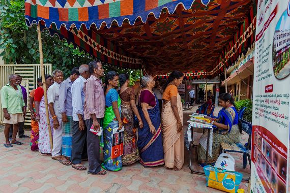 Frauen in Saris und Männer stehen unter einem bunten Stoffbaldachin Schlange.