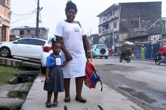Eine Frau in weißem Kittel hält ein kleines Mädchen mit Schulranzen an der Hand. Sie stehen auf dem Bürgersteig.