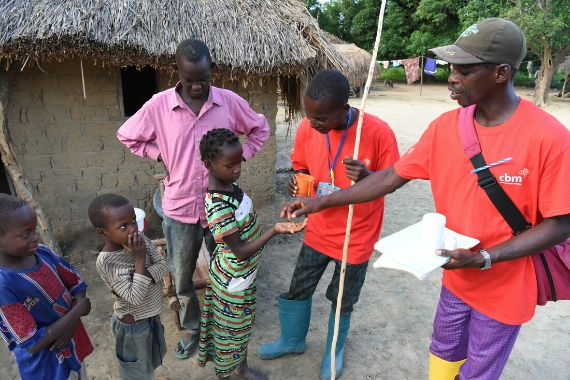 Ein Mann im roten T-Shirt legt einem Kind eine Tablette auf die hingestreckte Handfläche.
