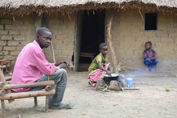 Ein etwa 11-jähriges Mädchen kocht an einem offenen Feuer vor einer Hütte, vor der ein Junge hockt. Ein Mann sitzt in der Nähe des Feuers auf einem aus Ästen gebauten Stuhl.