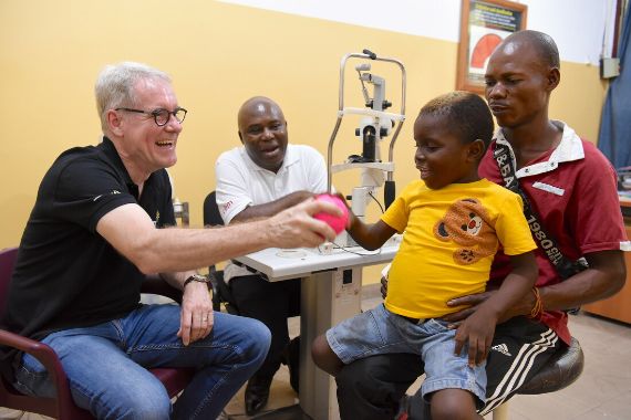 Ein Mann reicht einem Kind, das auf dem Schoß eines Mannes sitzt, einen bunten Ball. Das Kind nimmt den Ball entgegen, es lacht.