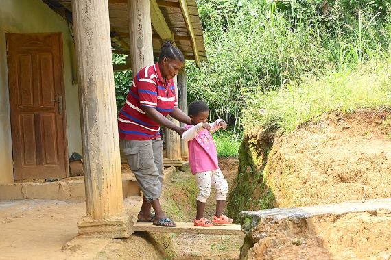Eine Frau und ein Kind gehen über ein Brett, das über einem kleinen Graben vor einem Haus liegt. Die Frau hält das Kind an den Oberarmen fest.