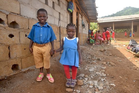 Ein etwa sechsjähriger Junge hält ein kleiners Mädchen an der Hand. Die Kinder lächeln. Sie stehen vor einer Mauer mit überstehendem Blechdach.