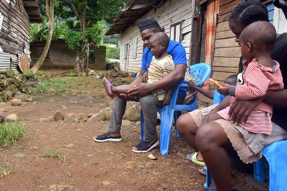 Ein Mann untersucht die Beine eines fünfjährigen Mädchens im Freien vor einer Hütte. Daneben sitzt eine Frau mit einem Baby auf dem Schoß.