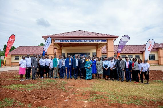 Vor einem eingeschossigen Gebäude mit offener Vorhalle steht eine große Gruppe festlich gekleideter Menschen.