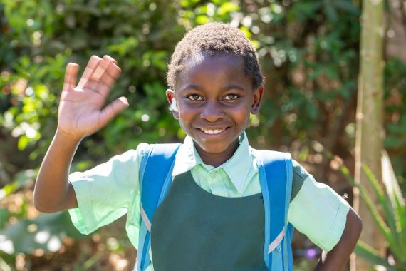 Ein Mädchen in Schuluniform mit zartgrüner Bluse und dunkelgrünem Trägerrock winkt lachend.