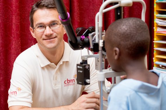 Ein Mann im weißen Polohemd mit CBM-Logo sitzt vor einem Augenuntersuchungsgerät, durch das ein etwa zehnjähriger Junge schaut.
