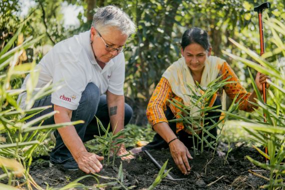 Eine Frau im Sari und eine Frau mit CBM-Polohemd sitzen in einem Beet mit noch kleinen Pfanzen.