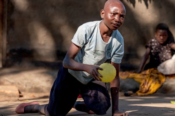 Junge aus Malawi kniet auf dem Boden und hält einen gelben Ball in seinen Händen