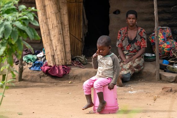 Ein kleiner Junge sitzt auf einem Stapel umgedrehter rosa Plastikeimer. Hinter ihm sitzt eine Frau vor einer Hütte.