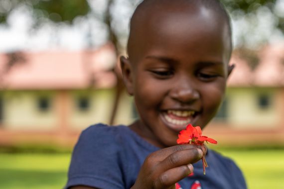 Kleines Mädchen aus Uganda hält lächelnd eine rote Blume in der Hand.