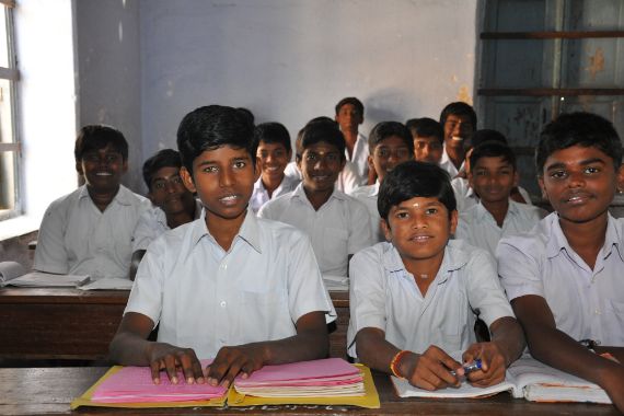 Ein blinder indischer Junge sitzt mit anderen Kindern in einem Klassenzimmer, vor ihm auf dem Tisch ein aufgeschlagenes Heft.