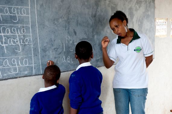 Eine afrikanische Frau steht an einer Schultafel und spricht in Gebärdensprache mit zwei Schulkindern.