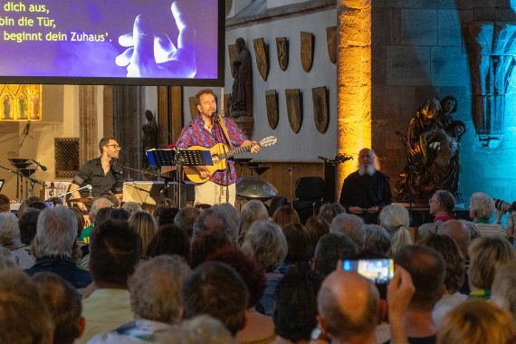 Ein Mann im karierten Hemd mit Gitarre steht in einem Kirchenraum vor Zuschauern.