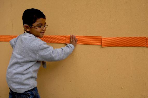 Ein kleiner Junge mit Brille tastet sich an einem taktilen Leitsystem an der Wand entlang.