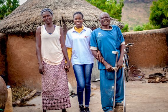 Vor einer Rundhütte: Eine Frau im T-Shirt mit CBM Logo legt die Arme um die Schultern einer weiteren Frau und eines Mannes mit Unteramrstützen.