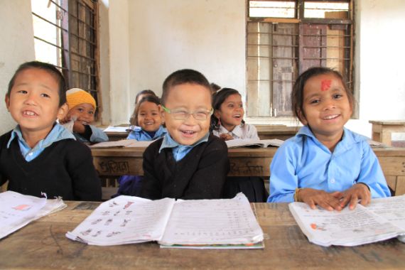 Ein kleiner Junge mit Brill sitzt neben zwei anderen Kindern lachend an einem Schulpult.