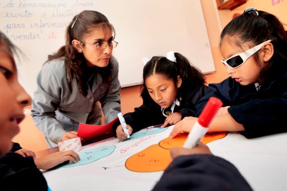 Eine Frau beugt sich über einen Tisch an dem mehrere Mädchen sitzen und etwas Schreiben. Eines der Mädchen trägt eine Brille mit dunklen Gläsern.