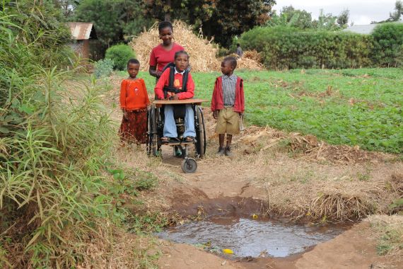 Ein Mädchen im Rollstuhl wird von einer Frau über einen Feldweg geschoben, in dem ein großes Wasserloch klafft.