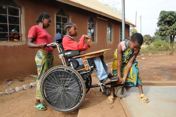Ein Mädchen im Rollstuhl wird von zwei Frauen über eine Stufe gehoben.