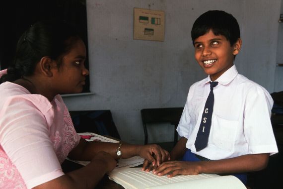 Ein blinder Junge in Schuluniform sitzt mit einer Frau an einem Tisch und tastet mit den Fingern über ein offenes Heft.