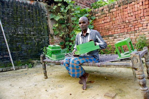 Ein alter Mann sitzt auf einer Bank und hält ein Modell für Kompostgruben in der Hand.