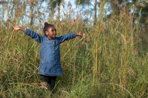 Ein Mädchen mit Hörgerät steht mit ausgebreiteten armen in hohem Gras