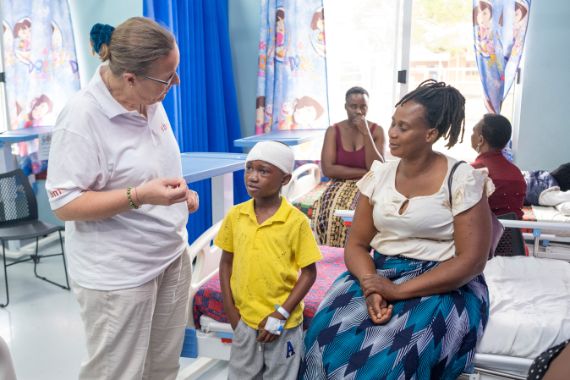 Eine Frau im weißen Polohemd steht vor einem kleinen Jungen mit Kopfverband. Neben dem Kind sitzt eine Frau in bunter Kleidung auf einem Krankenhausbett.