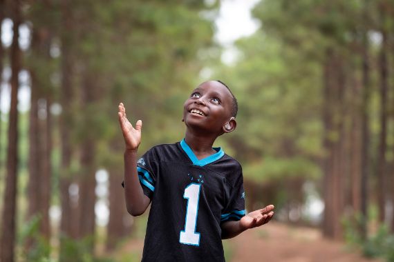 Ein lachender Junge mit Hörgeräten steht zwischen Bäumen und hält die Hände hoch.