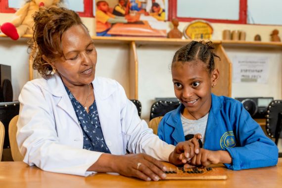 Ein junges Mädchen sitze mit einer Frau an einem Tisch. Vor ihr liegt eine Braille-Tafel, über die das Mädchen mit den Fingern tastet.