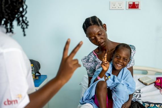 Jemand hält zwei Finger hoch und ein kleines Mädchen auf dem Schoß einer Frau macht es nach.