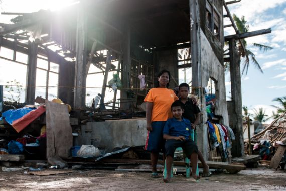 Eine Frau und zwei Kinder vor einer Hütte, deren Wände durch Taifun Hayian teils weggerissen sind und deren Dach beschädigt ist.