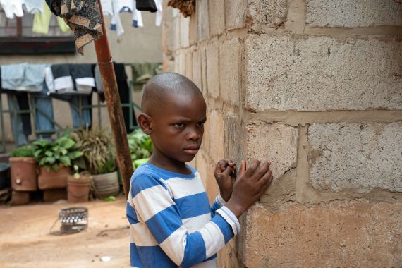 Ein etwa siebenjähriger Junge legt beide Hände an eine unverputzte Hauswand. Hinter ihm flattert Wäsche auf einer Leine.
