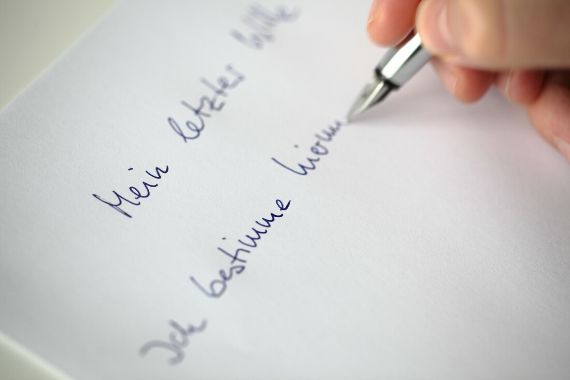 Eine Hand schreibt mit Tinte auf ein Blatt Papier &quot;Mein letzter Wille – Ich bestimme hiermit ...&quot;.