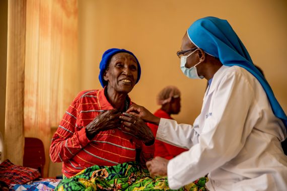 Eine Frau in Schwesterntracht sitzt am Bett einer alten Frau in bunter Kleidung. Beide lachen.