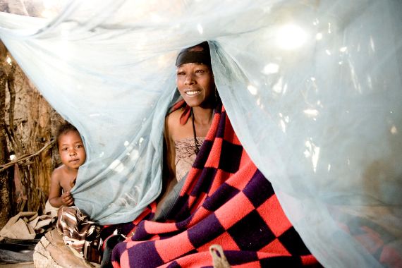 Eine in eine Decke eingehüllte Frau und ein Kleinkind sitzen auf einem Bett unter einem Moskitonetz.