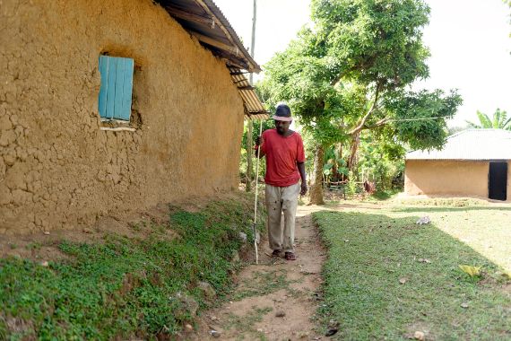 Ein afrikanischer Mann geht mit Stock auf dem Weg vor einer Hütte entlang.