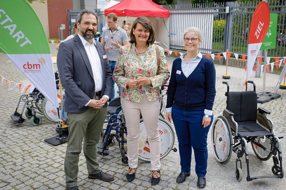 Auf dem Bild sind drei Personen abgebildet. Im Hintergrund sind Rollstühle und eine Fahne mit dem Logo der CBM.