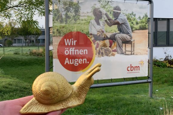 Auf dem Bild ist die Inklusionsschnecke vor einem CBM Plakat abgebildet. Auf dem Plakat steht: Wir öffnen Augen!