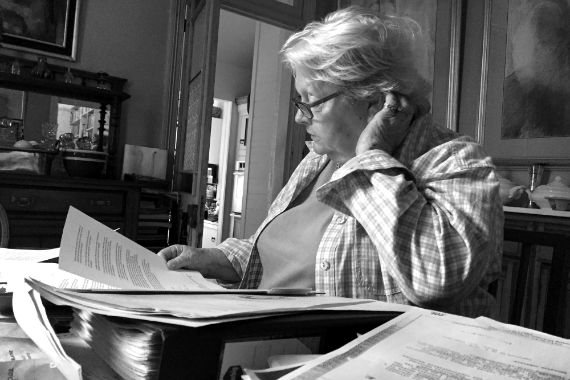 Ein Frau sitzt an einem Tisch, auf dem ein Ordner und einige Papierstapel liegen. Sie liest aufmerksam ein Dokument, das sie in der rechten Hand hält. Der linke Arm ist auf dem Tisch aufgestützt und sie greift hinter ihr Ohr. 