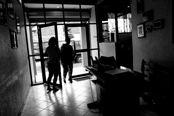 Eine Frau geht durch eine Glastür in ein Gebäude. Eine andere Frau hält ihr die Tür auf. Der Raum, in dem sie steht, sieht aus wie der Eingangsbereich eines Büros mit einem kleinen Schreibtisch als Empfang.