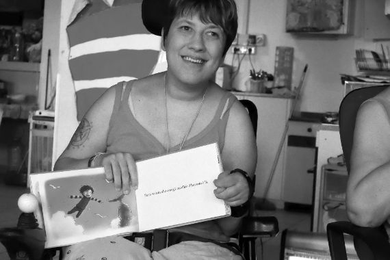 Eine lächelnde Frau sitzt in ihrem Rollstuhl und hält ein aufgeschlagenes Kinderbuch im Schoß.