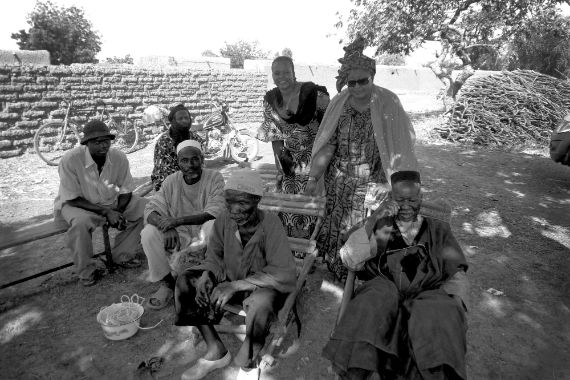 Zwei Frauen stehen in einem Garten, vor und neben ihnen sitzen fünf Männer auf Stühlen und Bänken. 
