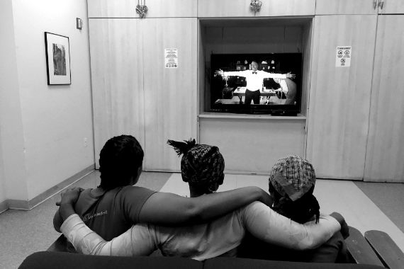 Rückenansicht dreier Frauen, die auf einem Sofa sitzen und Fernseher schauen: Sie legen sich gegenseitig die Arme um die Schultern. 