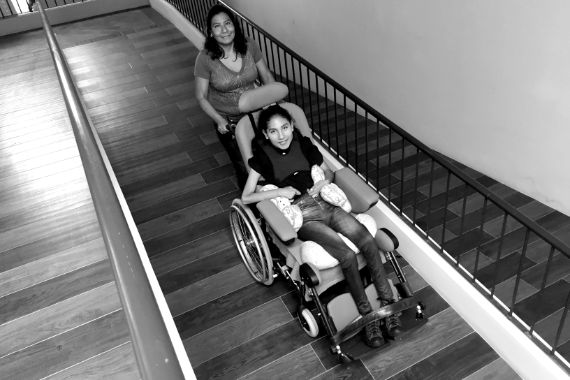 Eine junge Frau im Rollstuhl wird von einer anderen Frau eine Rampe hochgeschoben.