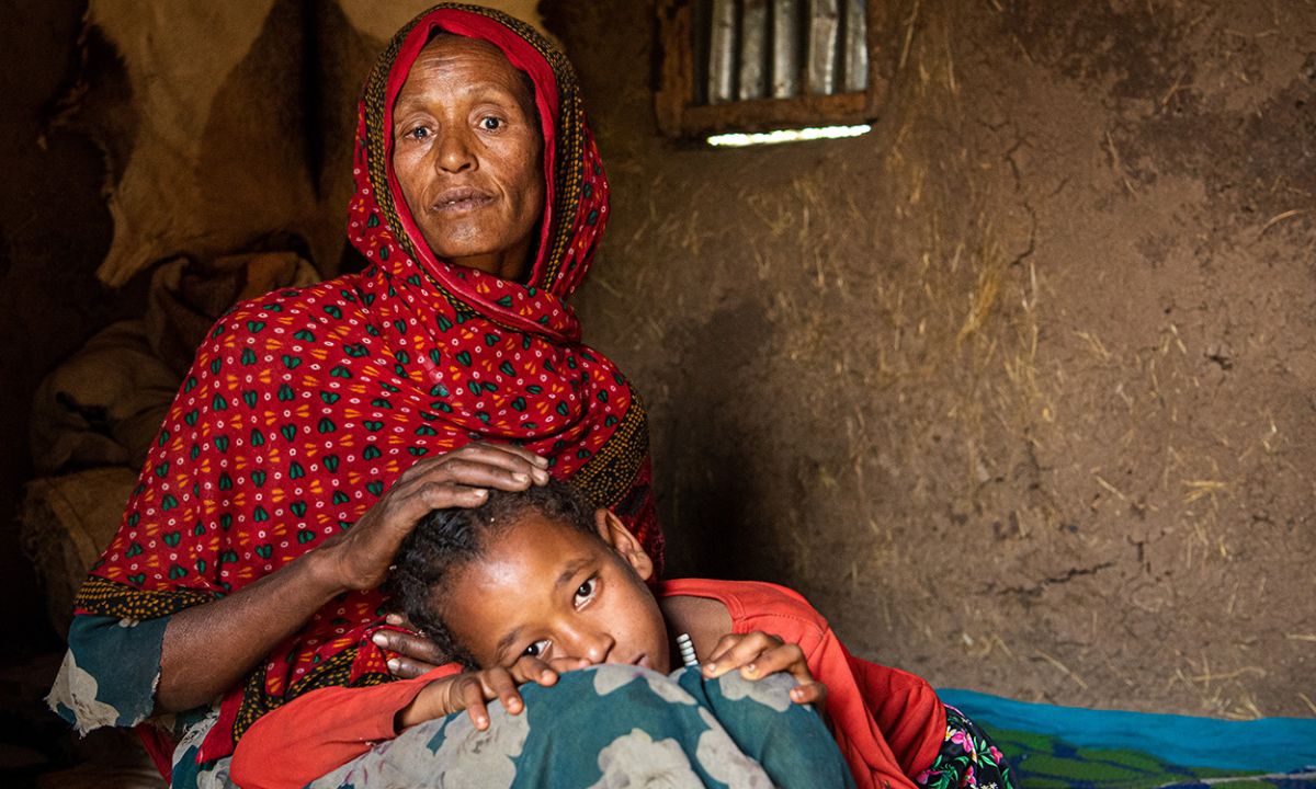 Eine Frau mit rotem Kopftuch sitzt in einer Hütte, ein Mädchen legt den Kopf in ihren Schoß.